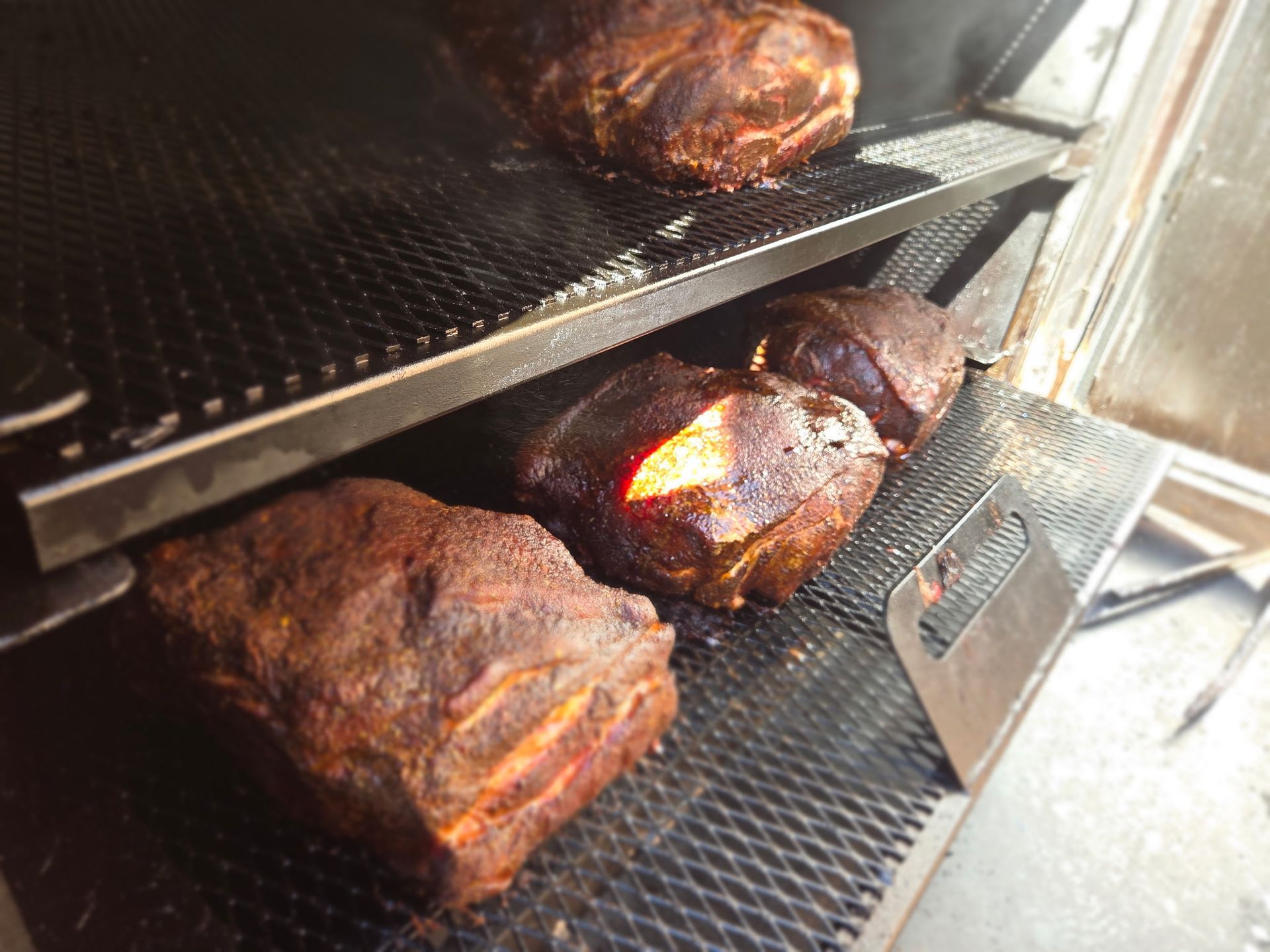 Smoked meat roasts on metal racks inside a smoker. The roasts are dark brown, with visible grill marks.
