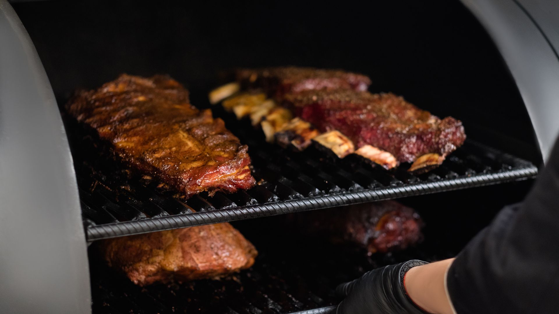 Smoked ribs and meat in a grill, person's hand with black glove, dark interior, food photography.