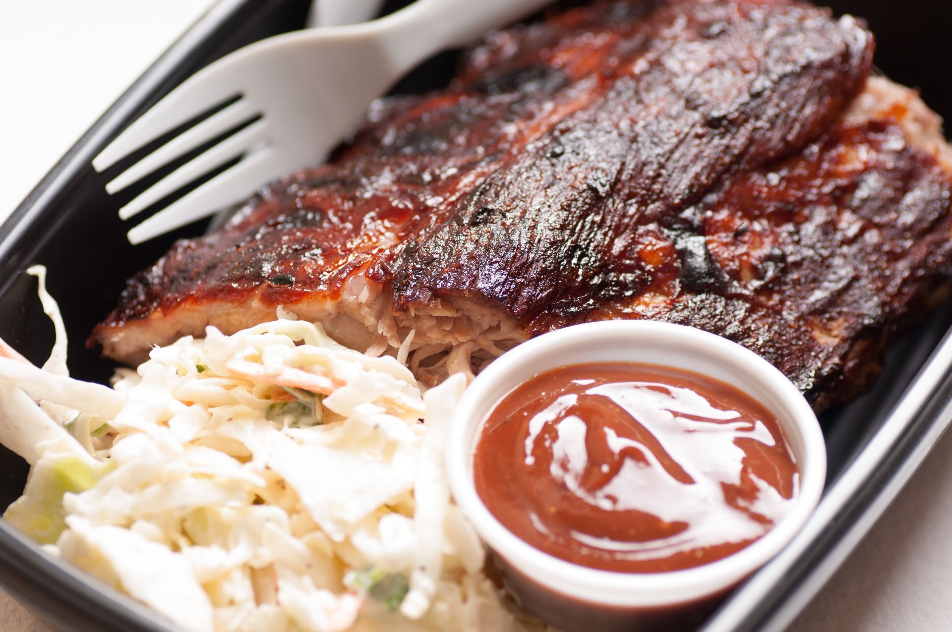 Barbecue ribs, coleslaw, and sauce in a black takeout container with plastic utensils.
