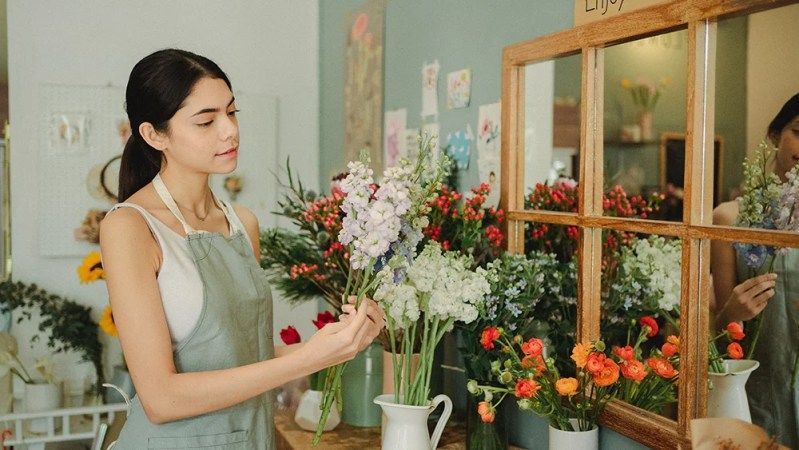 A woman is standing in a flower shop holding a vase of flowers.