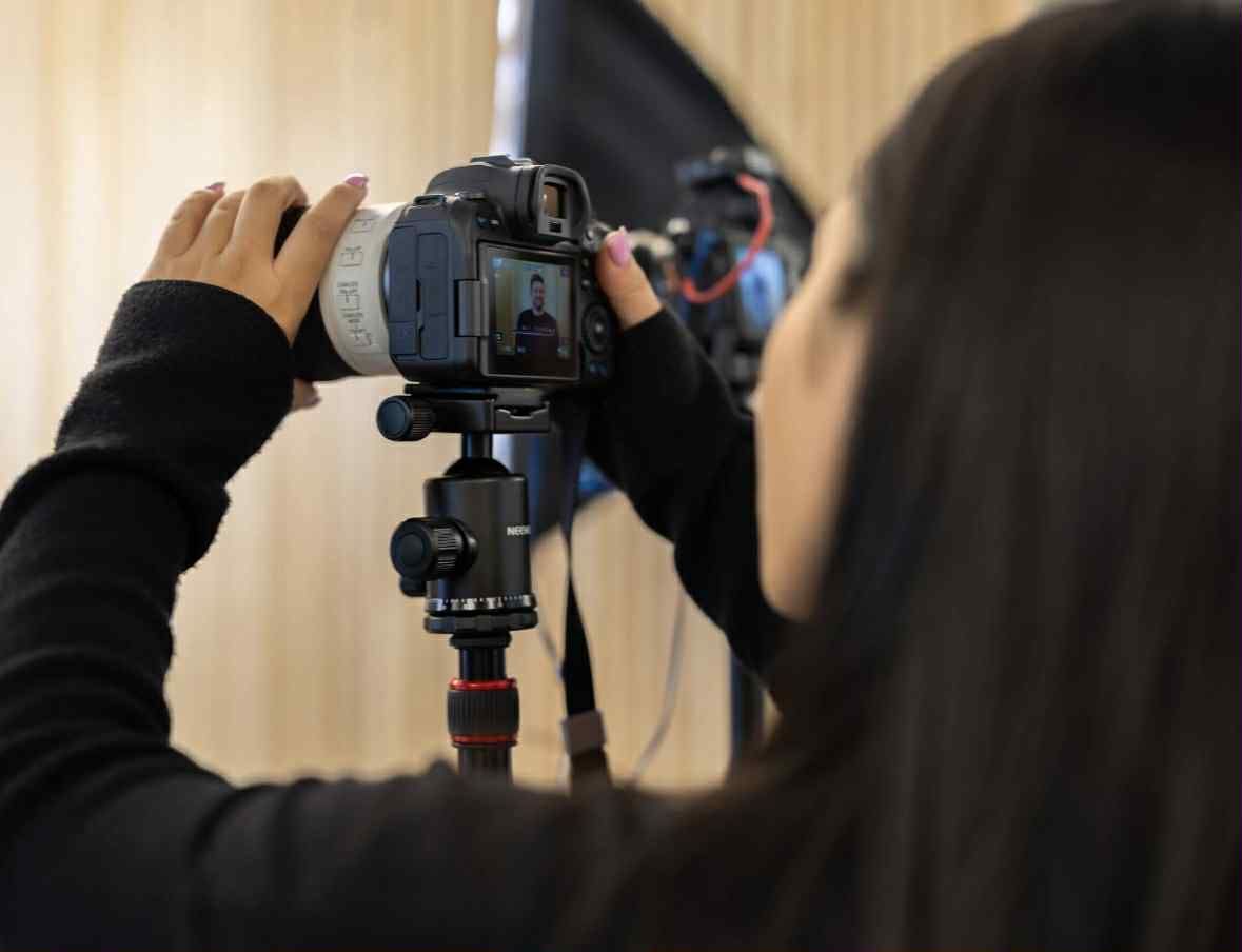 Woman looking through camera lens on a tripod, hands on the device. Studio setting. Woman looking through camera lens on a tripod, hands on the device. Studio setting.