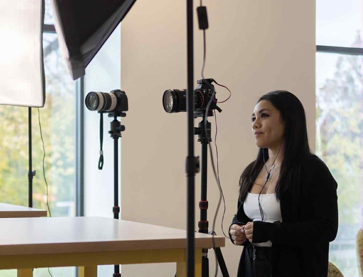 Woman in front of two cameras on tripods. Studio setup with lighting and tables by a window. Woman in front of two cameras on tripods. Studio setup with lighting and tables by a window.