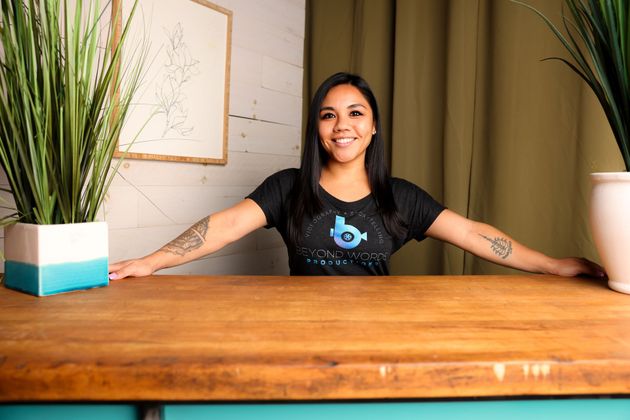 Woman smiling, arms outstretched, at a wooden table with plants on either side.