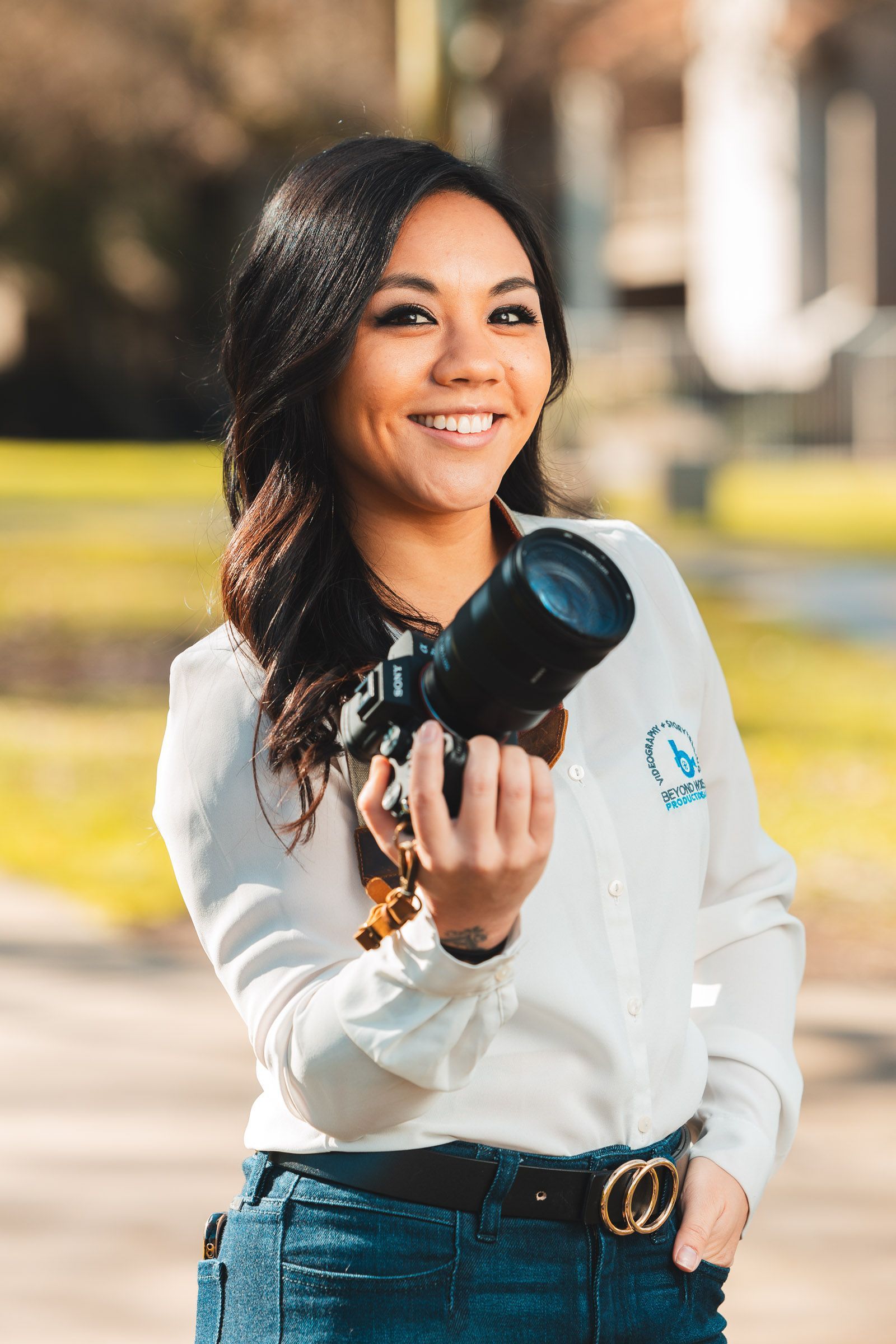 Woman with camera smiles, outdoors. Holding camera with large lens, white shirt, jeans. Blurred green background.