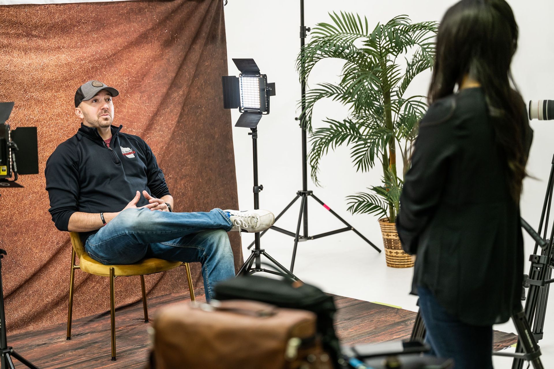 Man seated in a studio interview setup while another person stands in the foreground near lights and backdrop.