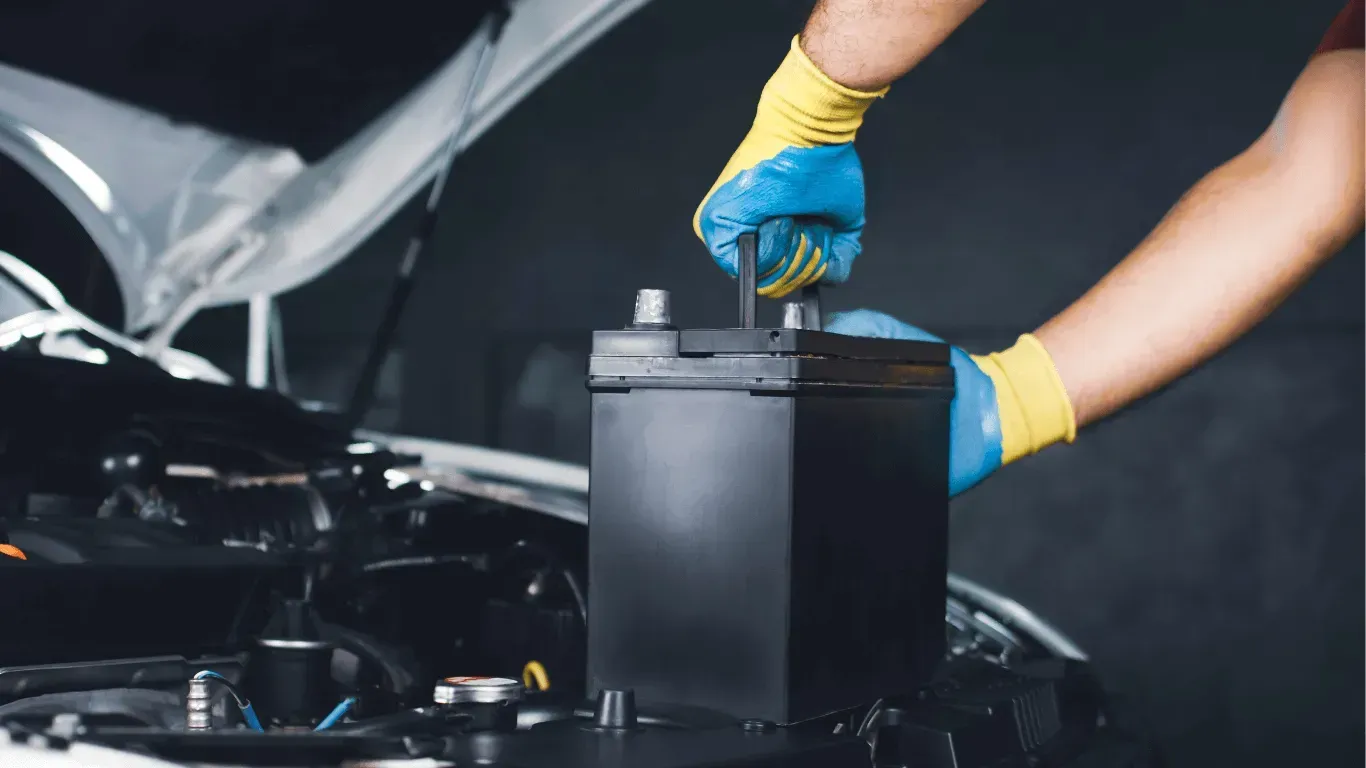 A mechanic wearing blue and yellow gloves lifts a black car battery from an open vehicle hood in a garage.