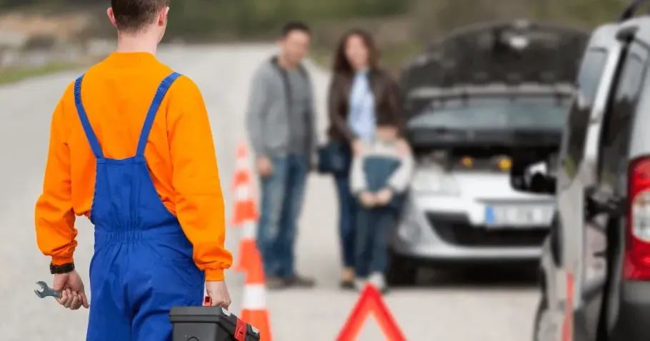 A mechanic in an orange shirt and blue overalls walks toward a family standing by a car with its hood raised on a road.