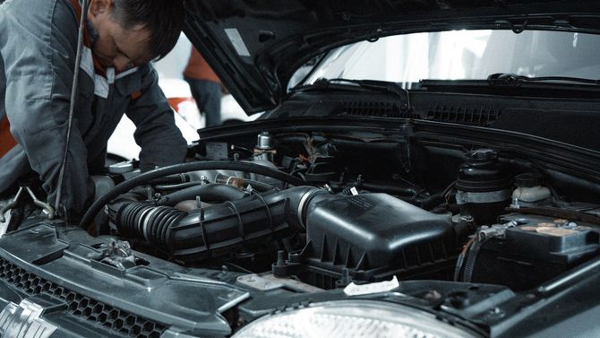A person in a gray jumpsuit leans over an open car hood, performing maintenance on the engine.