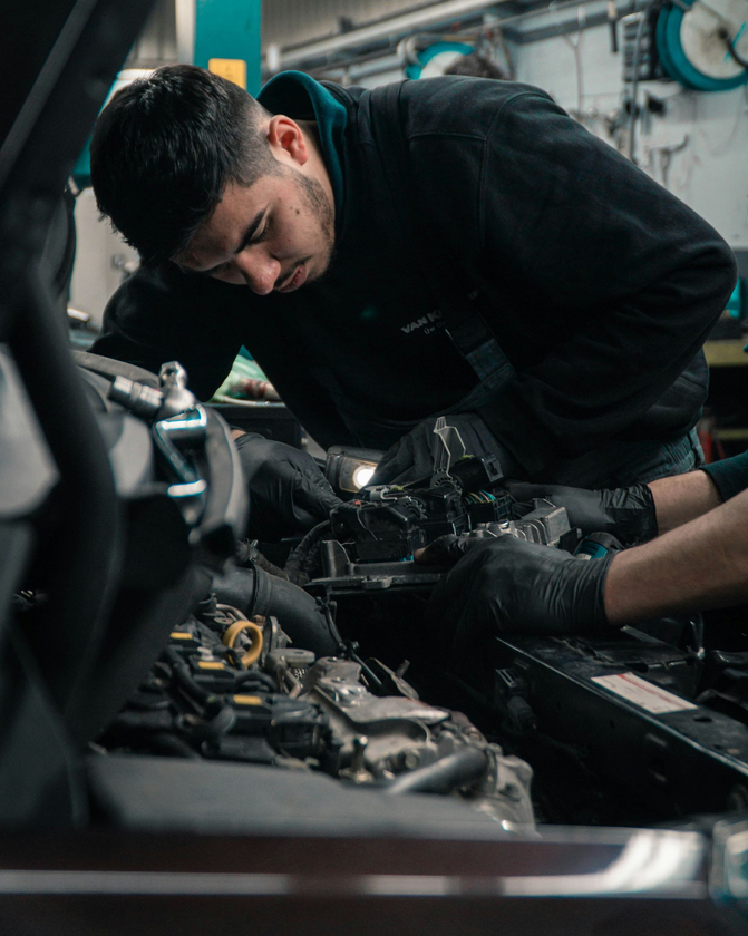 A mechanic wearing gloves works on an open car engine in an auto repair shop.