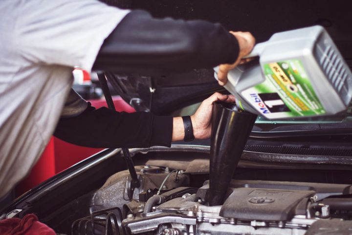 A mechanic pours Mobil 1 motor oil through a black funnel into a car engine.