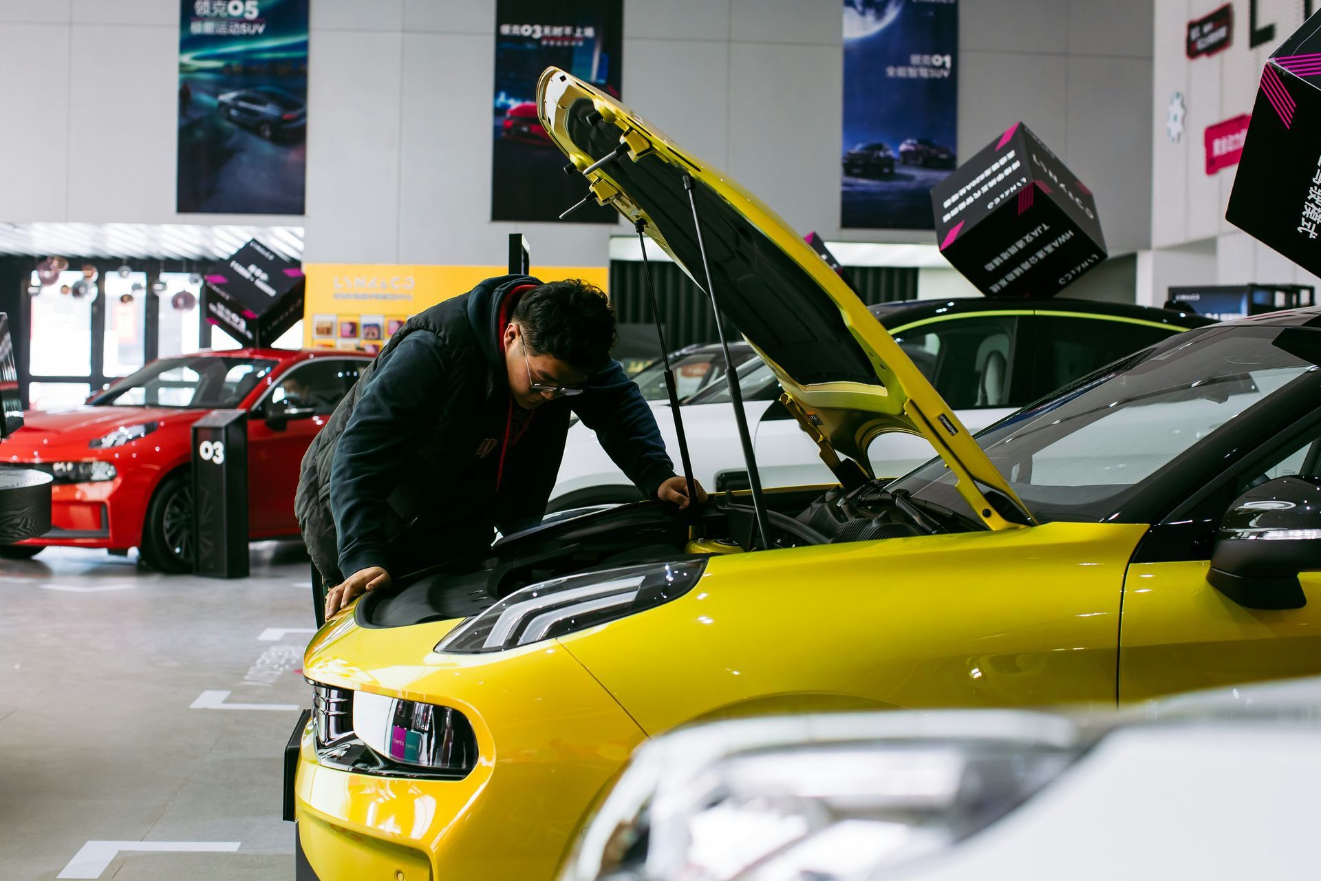 A person leans over the open hood of a yellow car inside a brightly lit car showroom with other vehicles on display.