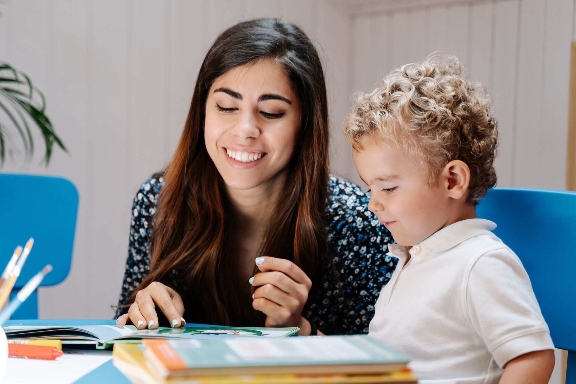 A woman helping a child read a colourful book at a table with pencils and stacked books nearby A woman helping a child read a colourful book at a table with pencils and stacked books nearby