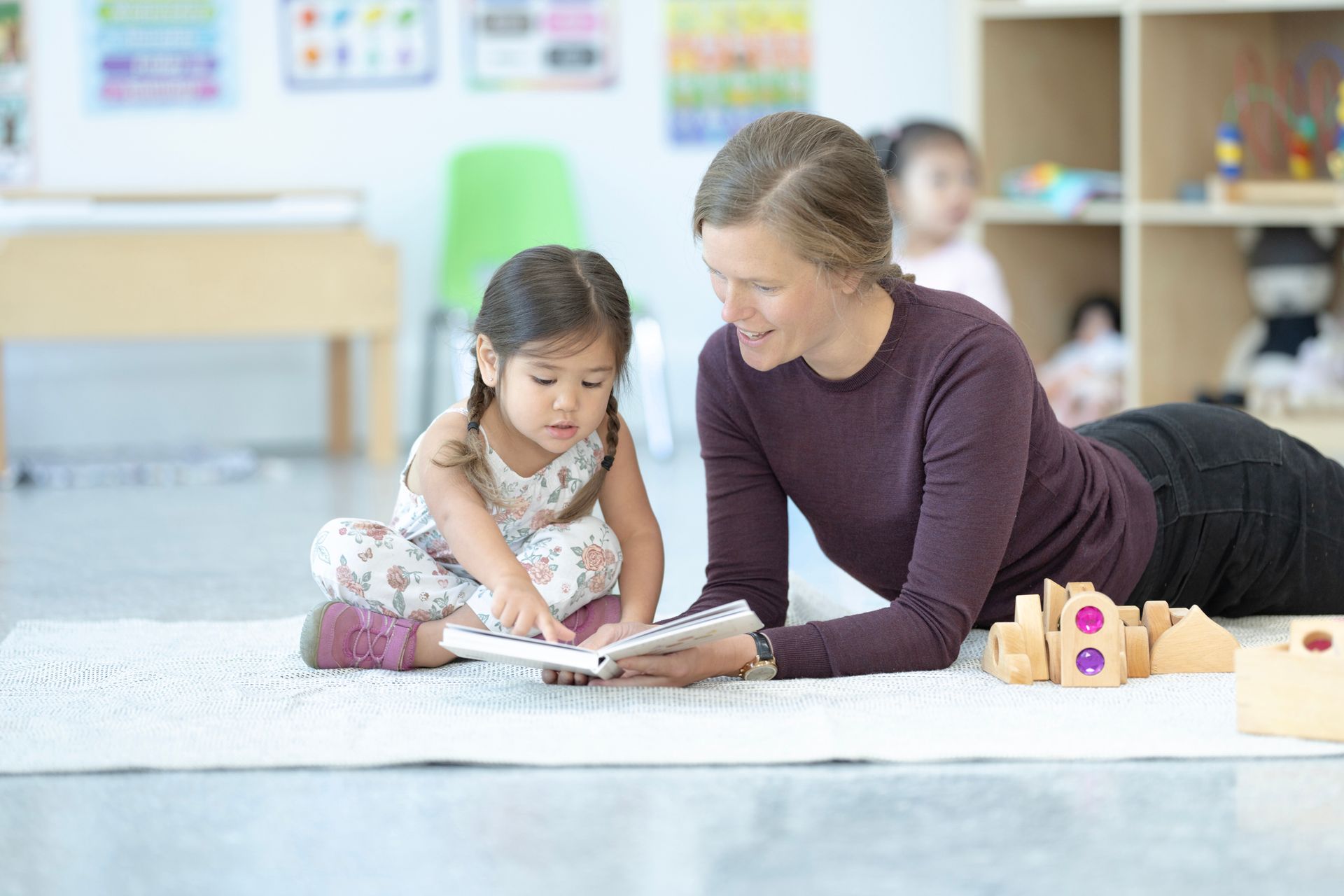 A woman lying on the floor with a child, reading a book together in a bright classroom A woman lying on the floor with a child, reading a book together in a bright classroom