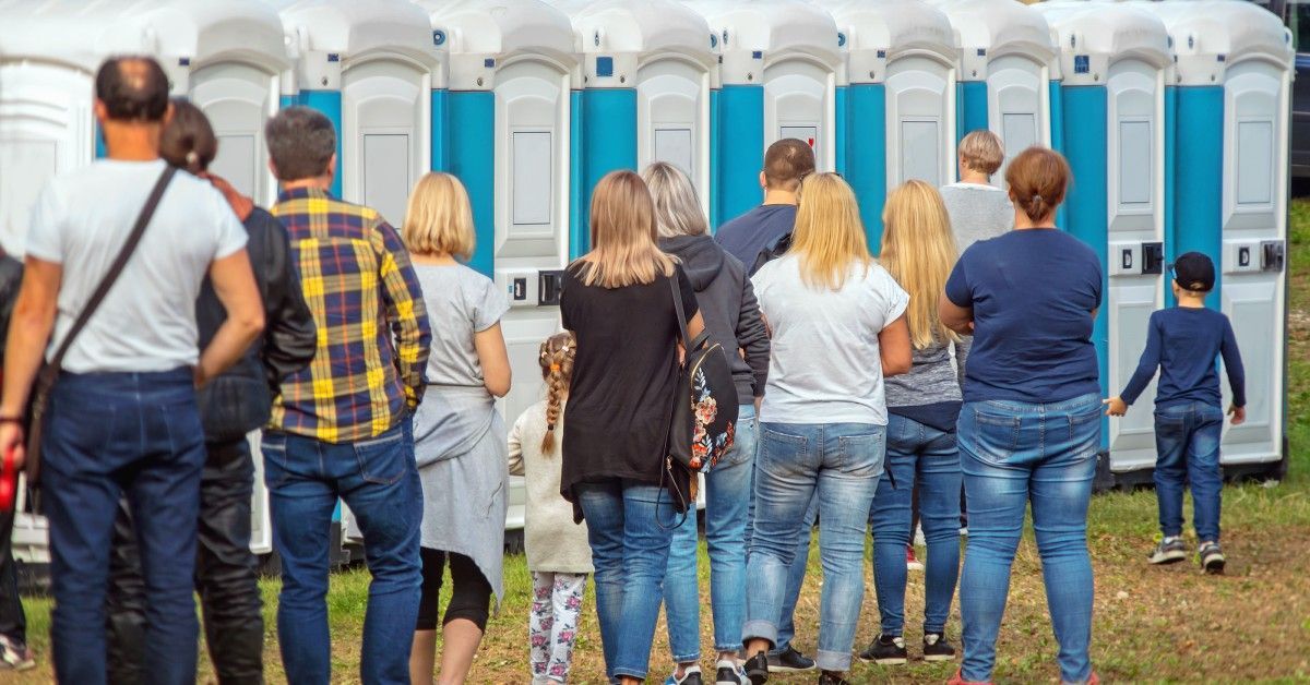 A line of people stand outside as they wait their turn in a portable toilet at an event. The units are blue and gray.