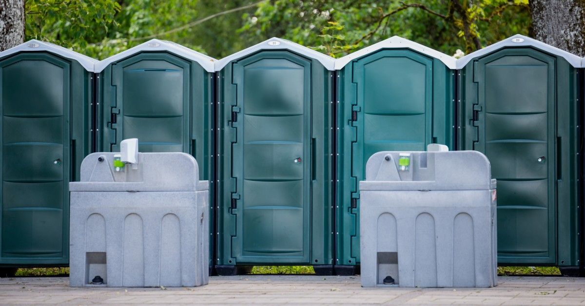A row of green portable toilets sit outside next to a tree line. There are two gray handwashing stations in front of them.