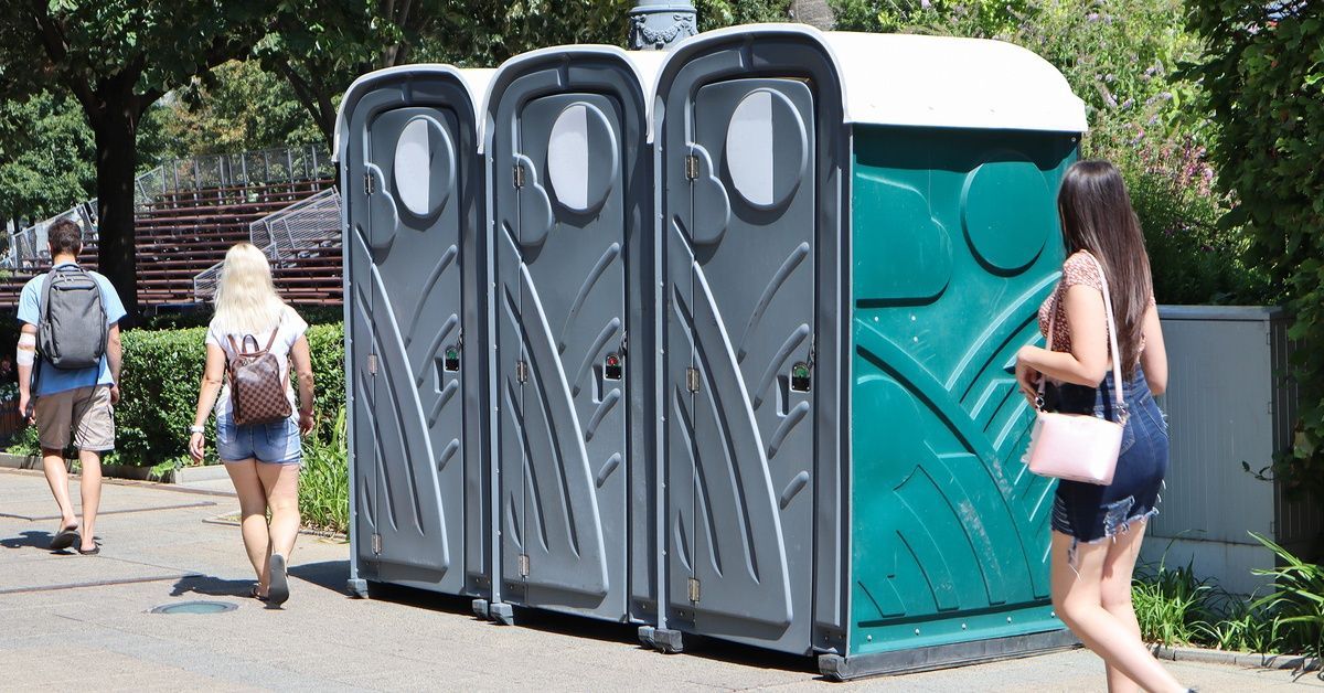 A group of three portable toilet units sitting on the street of a festival. Three attendees walk by the units.