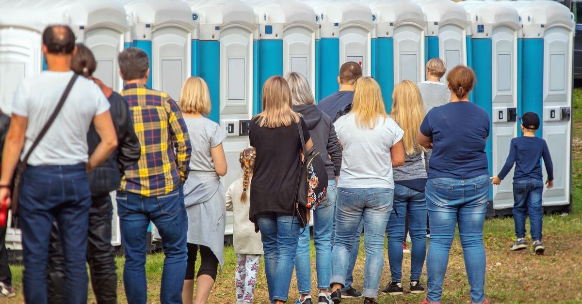A row of portable toilet units with a line of people standing, waiting to use one. The sun is shining outside.