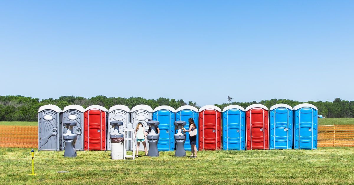 A row of gray, blue, and red portable toilet units sit in a field with a few users near the hand washing station.