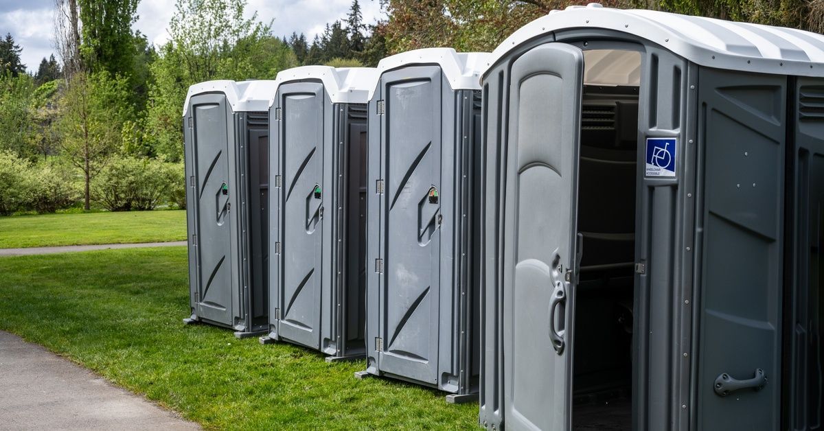 A row of portable toilets sit along the grass next to a paved sidewalk. The first unit on the right