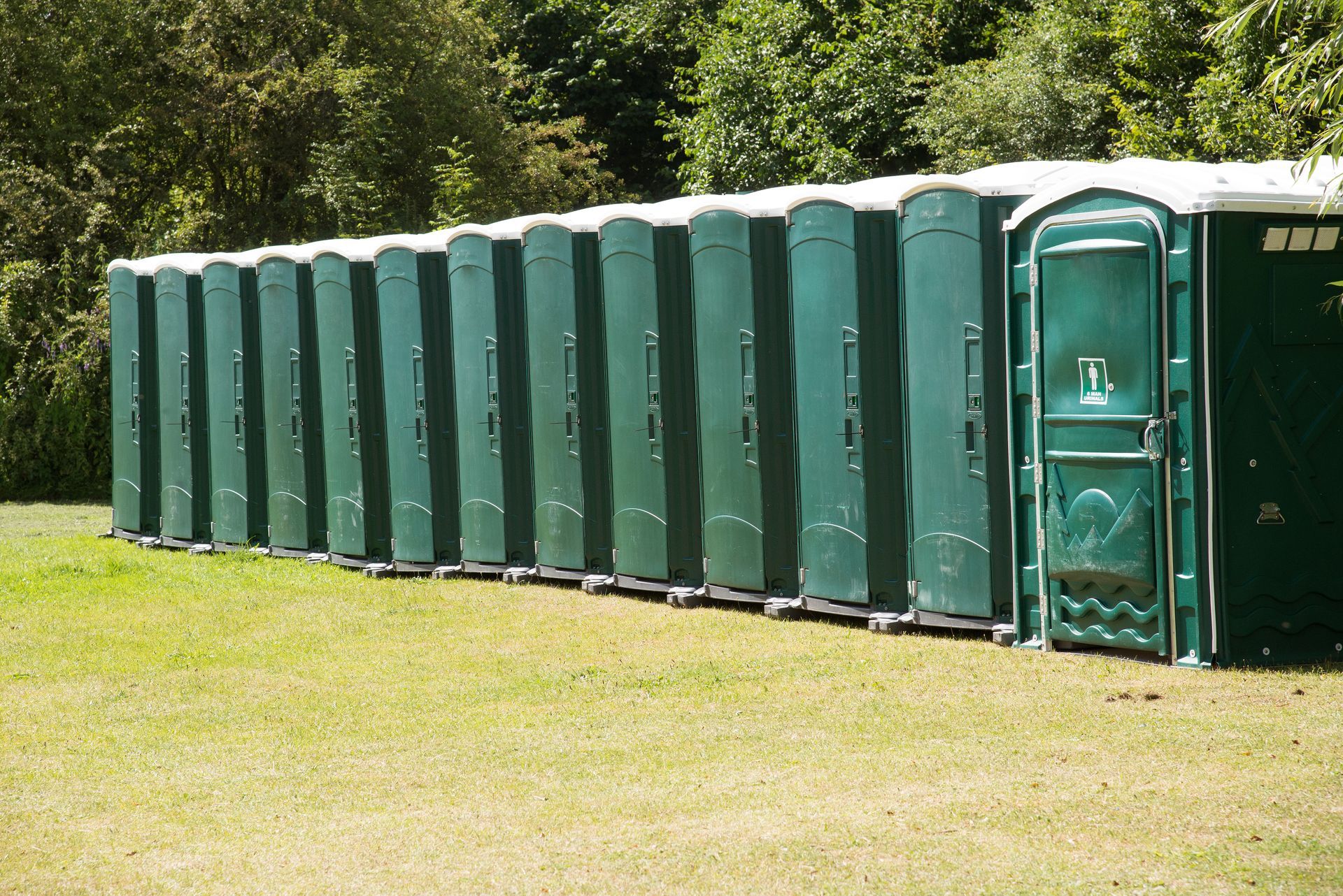 A row of green portable toilets lined up for an outdoor event. All the doors are shut and the grass is short.