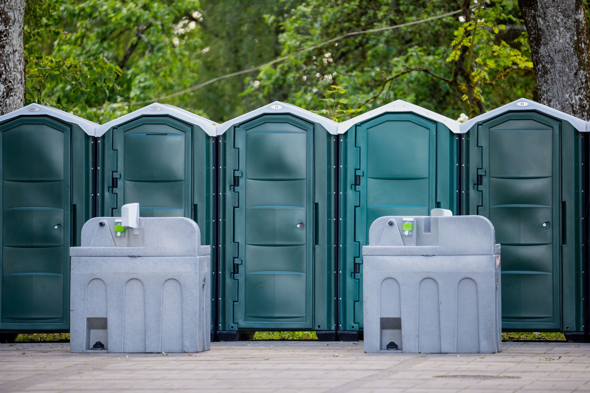 A row of green portable toilets sit outside at an event. There are two portable handwashing stations in front.