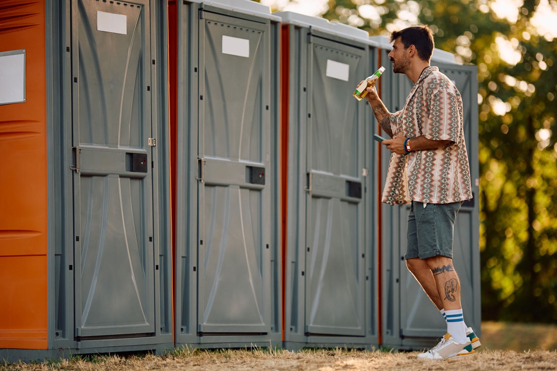 A young man drinks a beverage at an event as he waits in line for a portable toilet to open. He stan