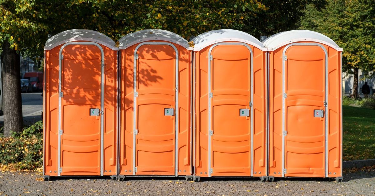 A row of four orange mobile toilet units sit in a park on a sunny day. The units are clean and three are unoccupied.