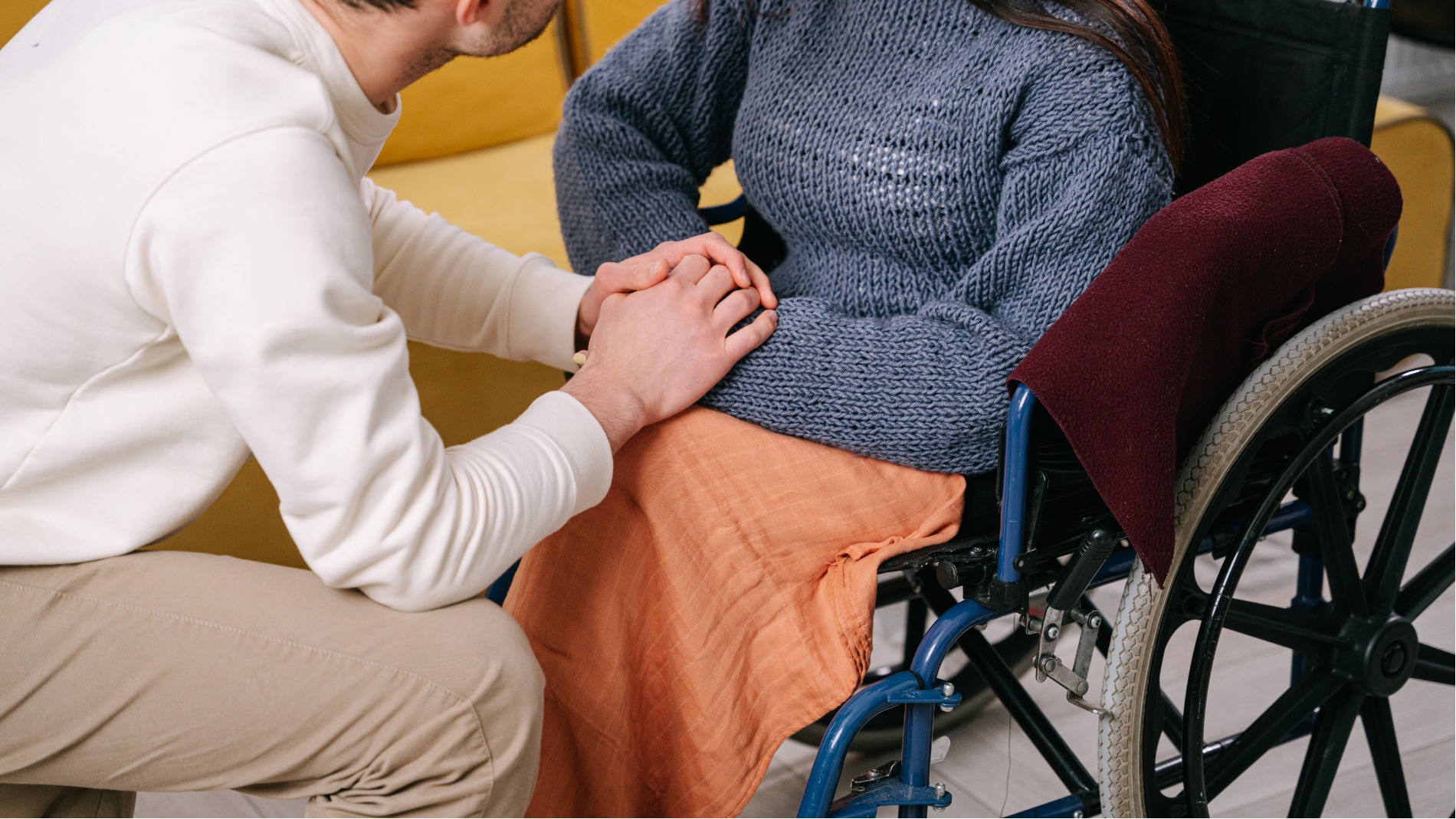 Person in a wheelchair receiving support from another person, with a hand on their shoulder and knee.
