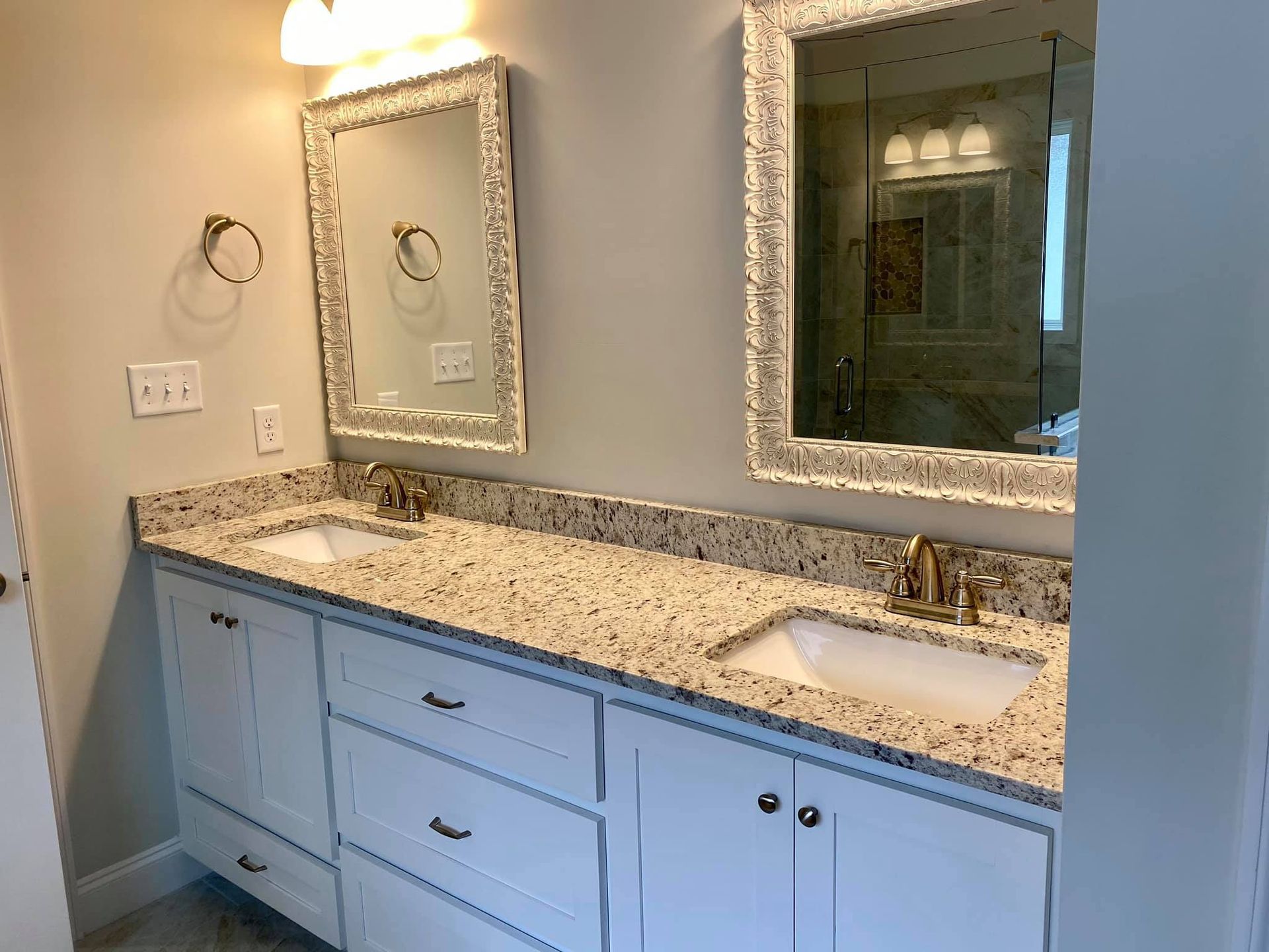 Bathroom with double sinks, white cabinets, granite countertop, ornate mirrors, and gold fixtures.