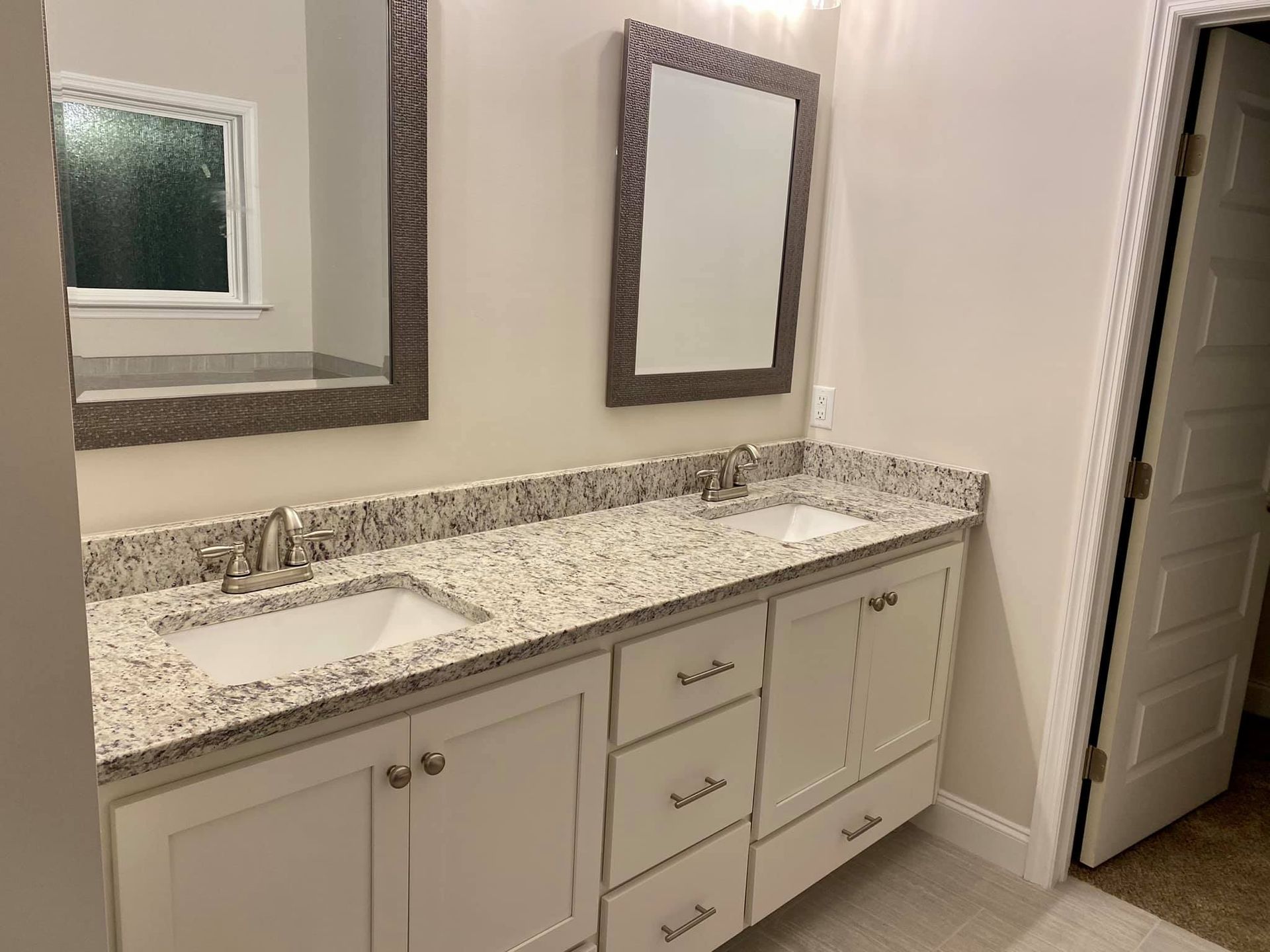 Bathroom with double vanity, granite countertop, two mirrors, white cabinets, and a partially open door.