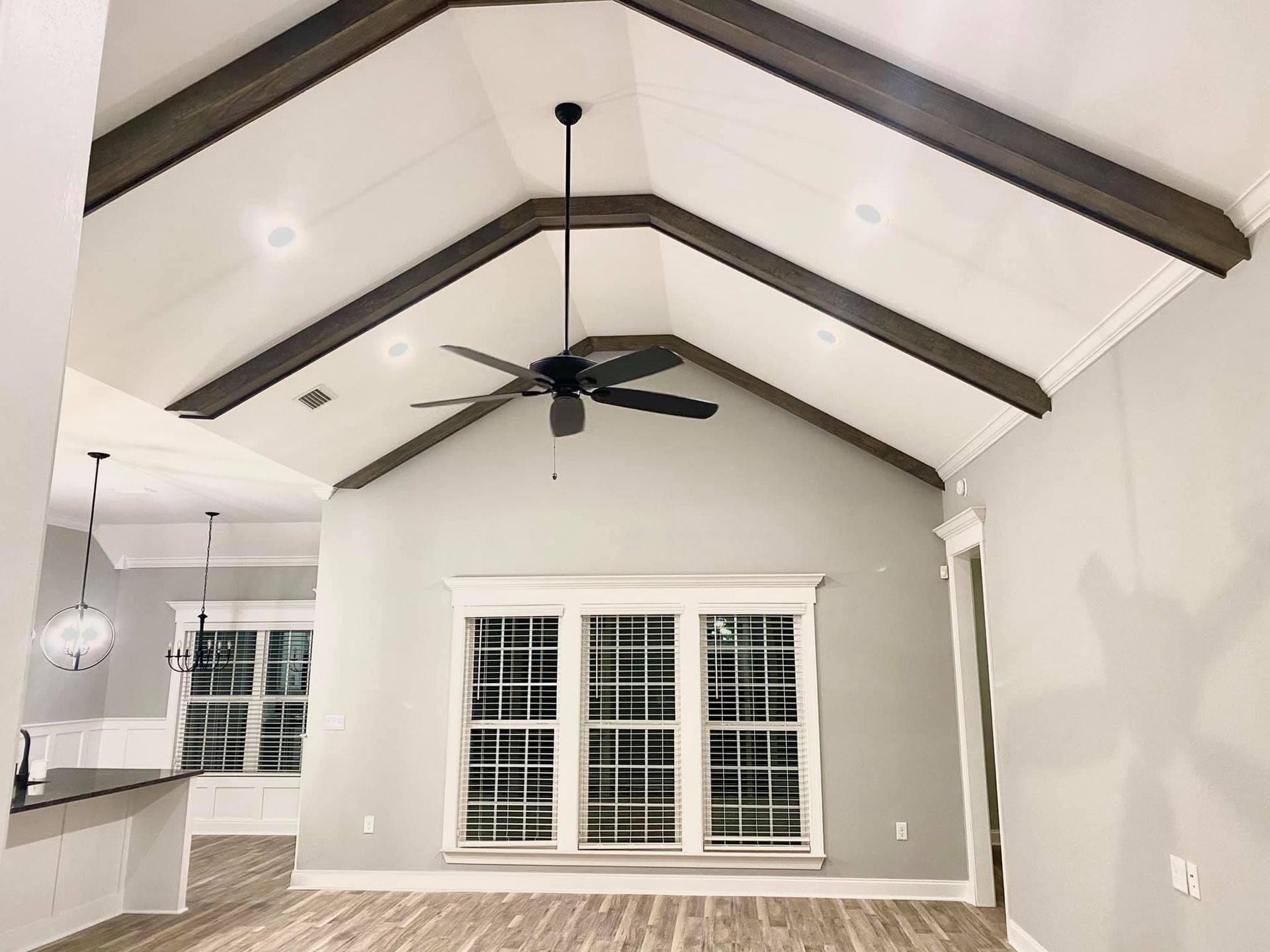 Vaulted ceiling with dark wood beams, a fan, and a large window in a gray-walled room.