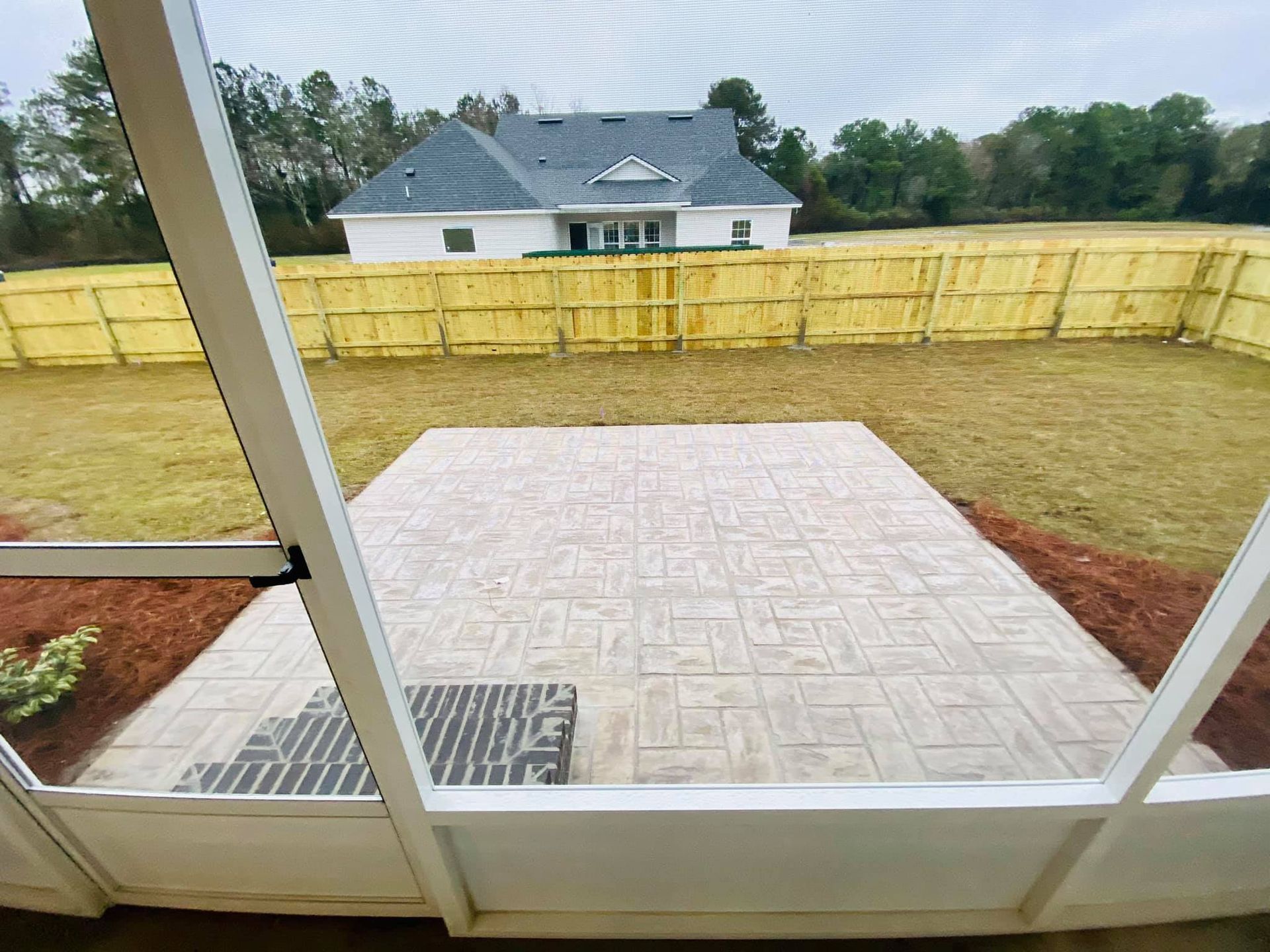 View from a screened porch of a patio, backyard, and a house with a wooden fence.