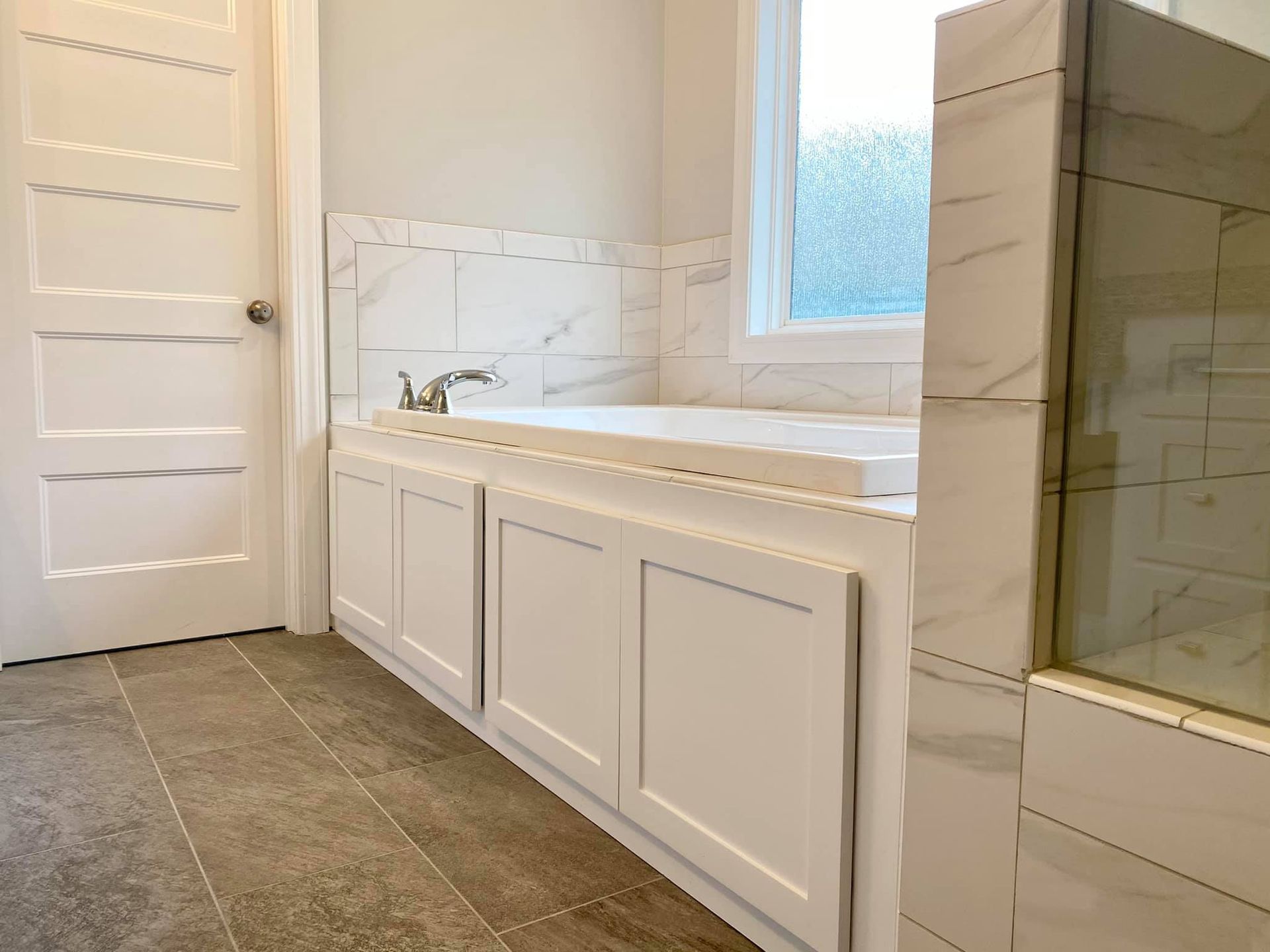Bathroom with white tub, cabinets, and marble tile surround. Door on left, window on right.