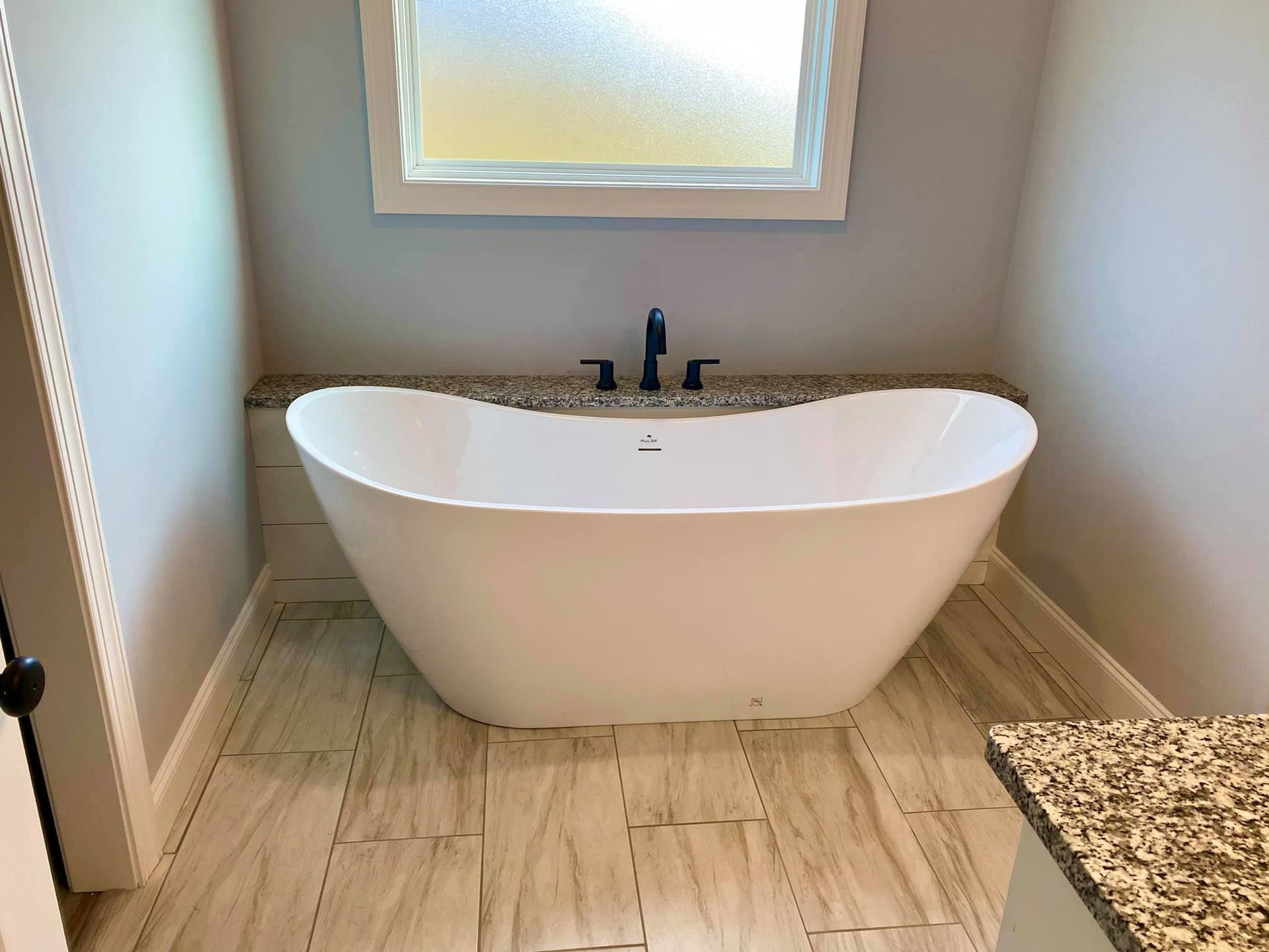 White freestanding bathtub in a bathroom, under a window, with light gray walls and stone tile floor.