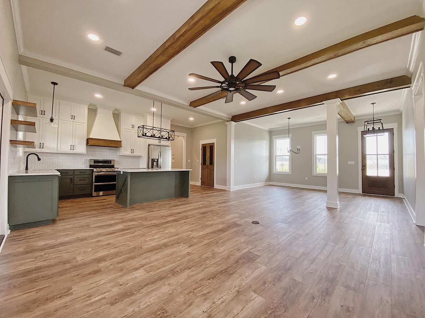 Open-concept kitchen and living area with wooden beams, light green cabinets, and a large fan.