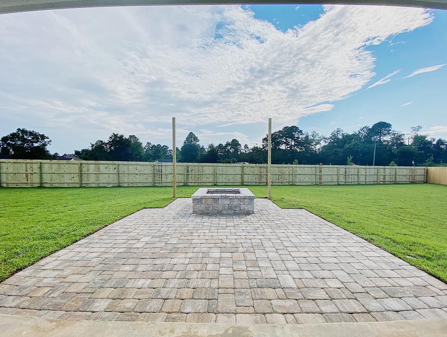 Backyard patio with fire pit, brick pavers, and green lawn, under a partly cloudy sky.