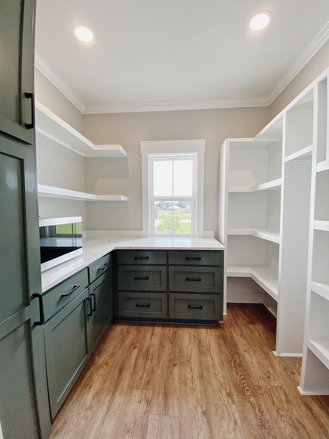 Pantry with gray-green cabinets, white shelves, and a window. Wood-look flooring and white walls.
