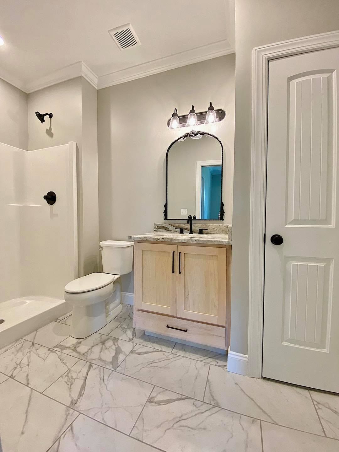 Bathroom with marble tile, light wood vanity, arched mirror, black fixtures, white shower, and door.