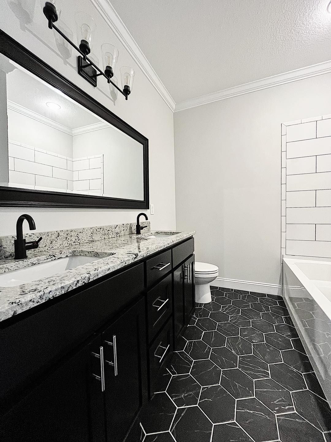 Black and white bathroom with dark hexagon floor tiles, double vanity, and a tub.