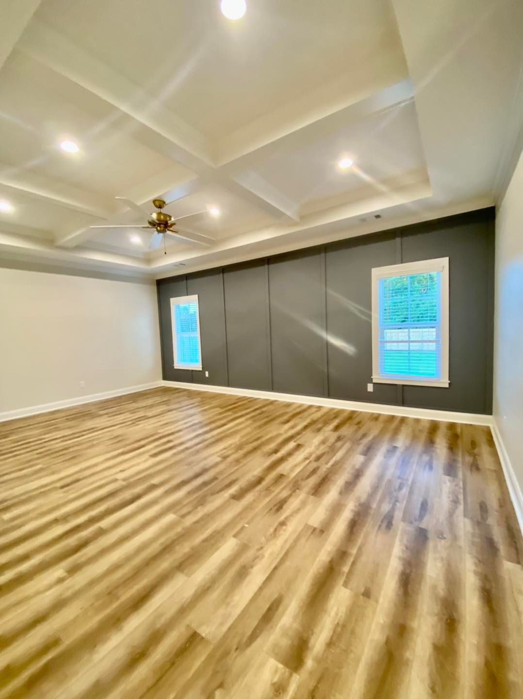 Empty room with wood-look floor, gray accent wall with two windows, and coffered ceiling with recessed lights.