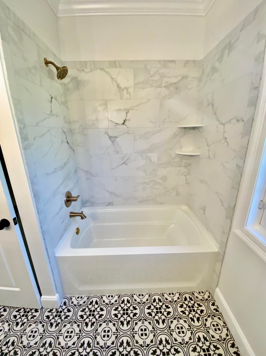 Bathroom with white tub, marble-look tiled walls, gold fixtures, and patterned black and white floor tiles.