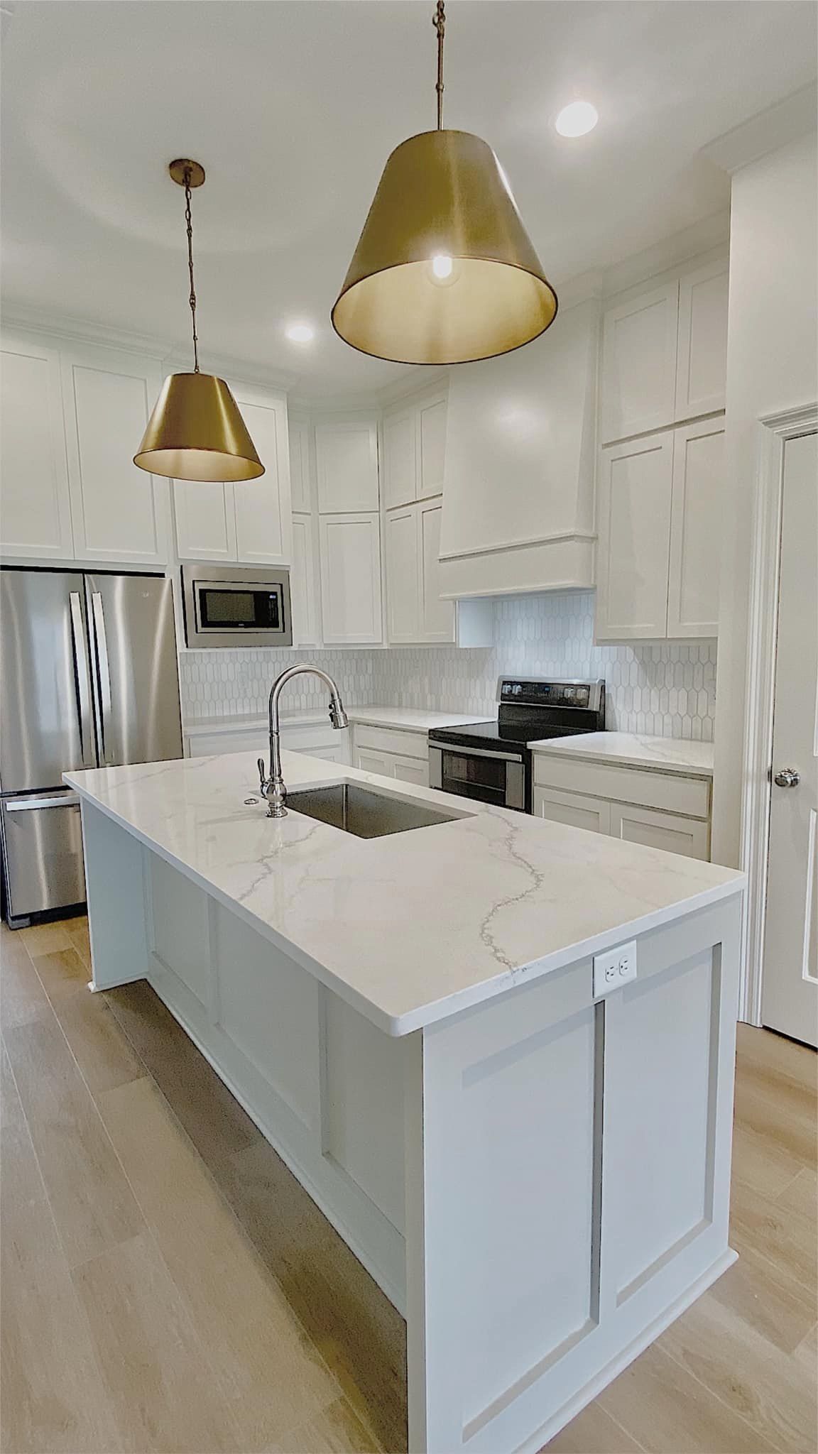 Modern white kitchen with a large island, brass pendant lights, and stainless steel appliances.