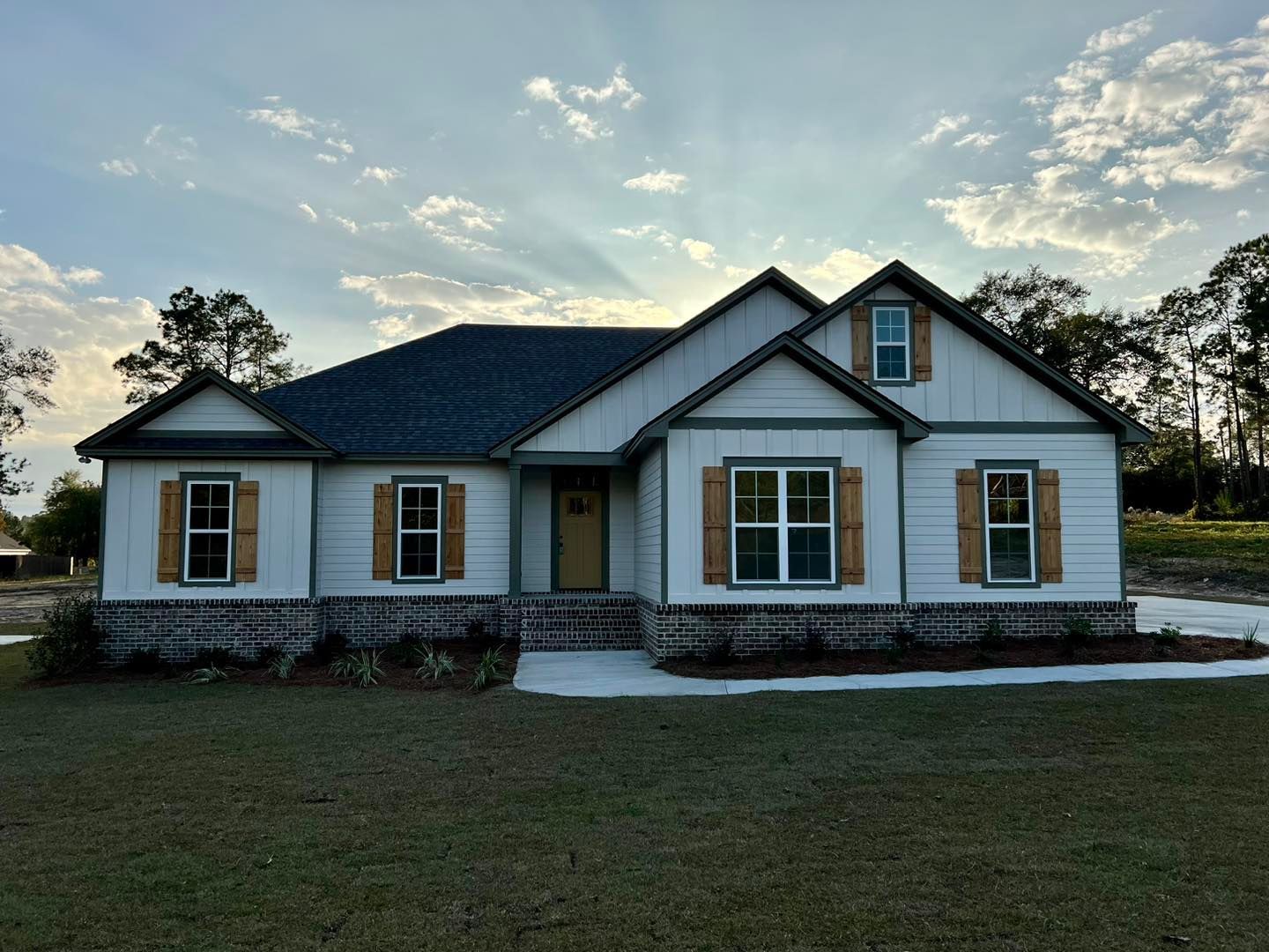 White house with brown shutters and a brick base under a cloudy sky.
