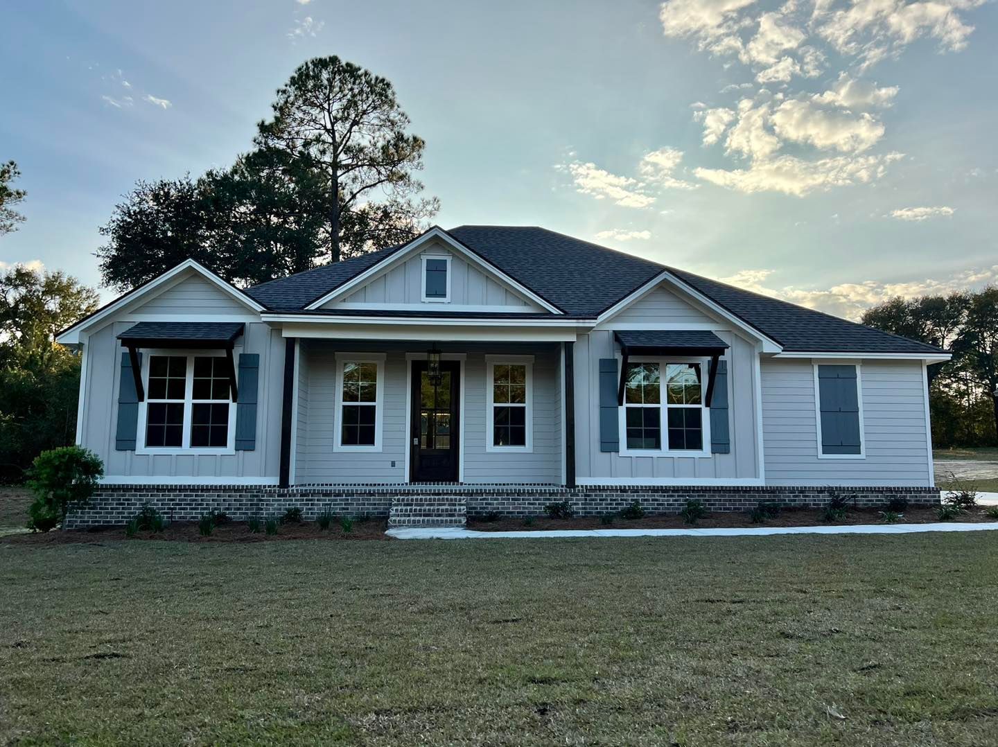 Light blue, single-story house with black awnings, dark roof, and blue shutters, sitting on a grassy lawn u