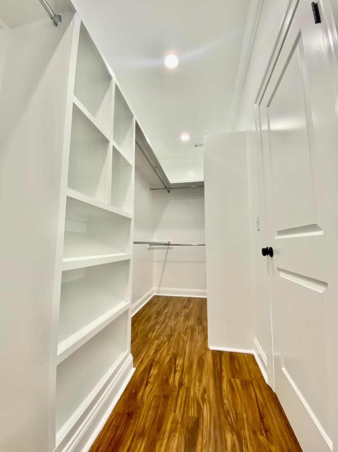 Bright white walk-in closet with shelves and rod. Wooden floor, door on right.
