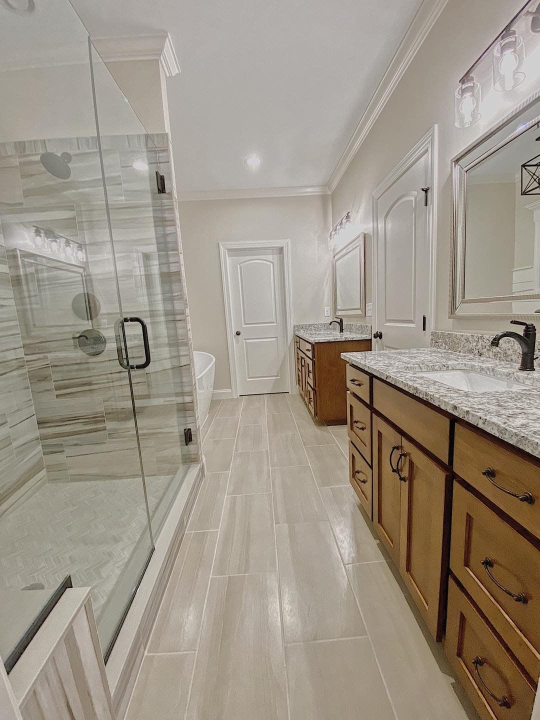 Bathroom with light tile floor, wood vanities, glass shower, and white door.