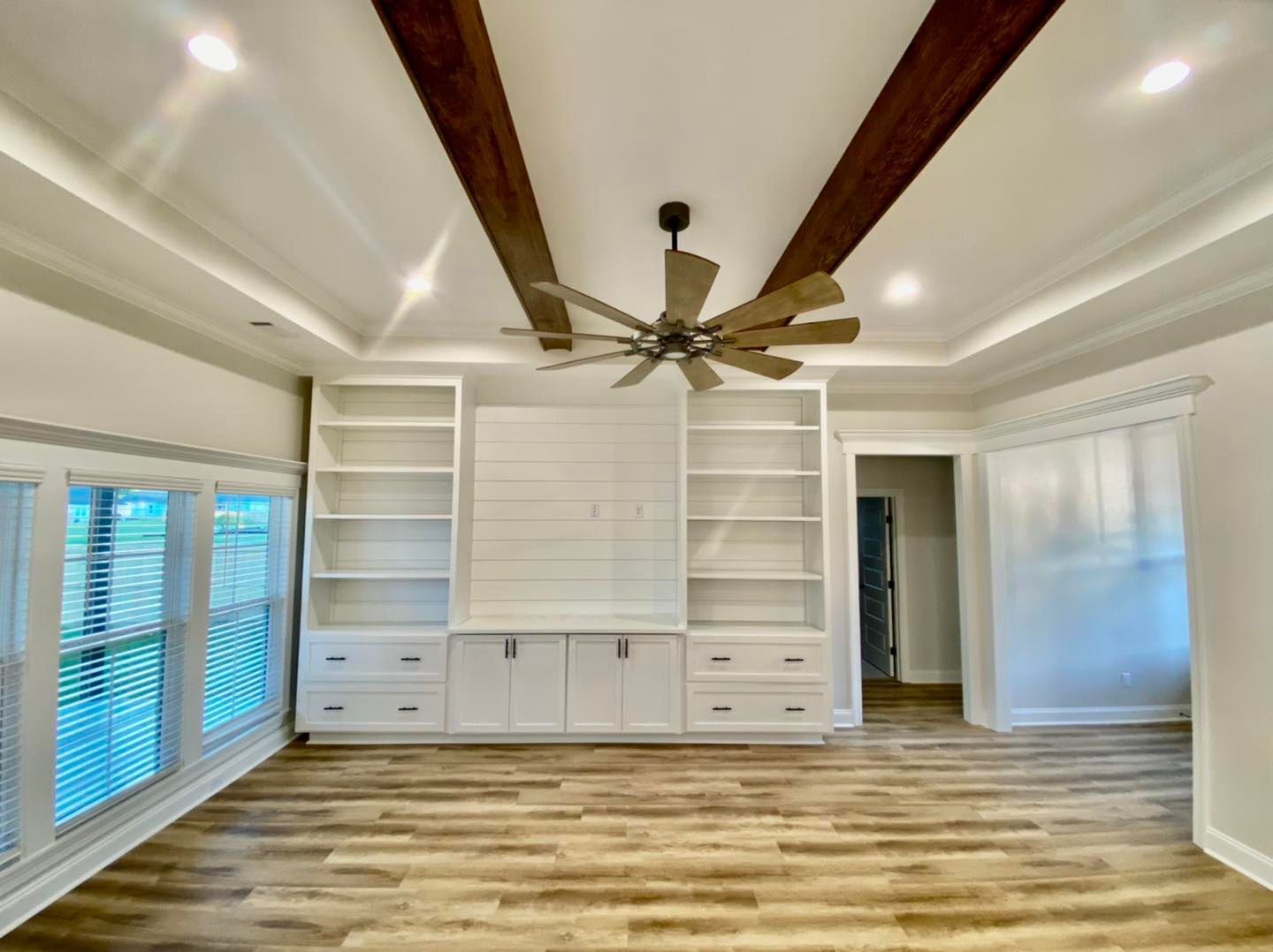 A bright living room with built-in white shelving, wooden beams, a ceiling fan, and hardwood floors.