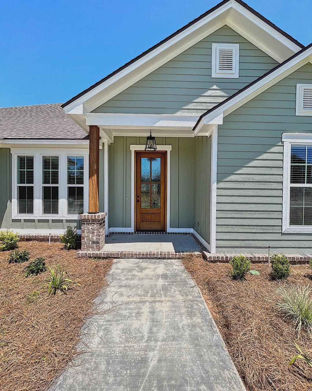 Green house with brown door, porch, and walkway. Brown wood chips and blue sky.