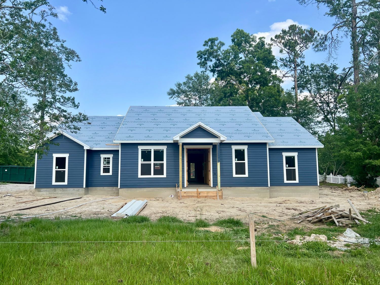 Blue-sided house under construction with a light blue roof, surrounded by green grass and trees.