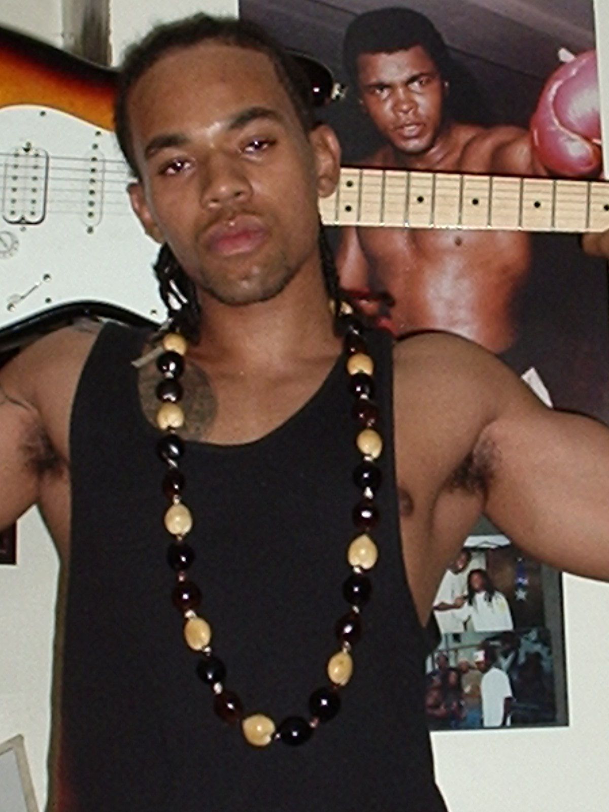 Robincito with Braids and necklace posing, electric guitar behind him, a Muhammad Ali poster in the background.